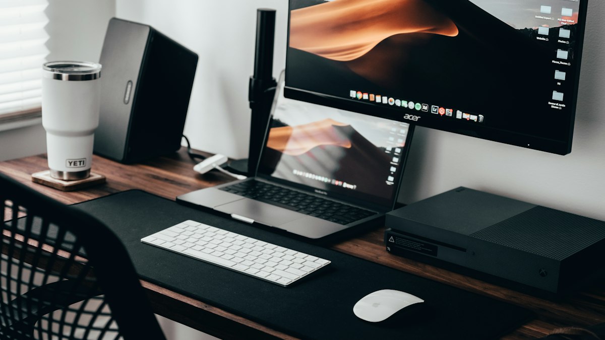 A desk with a laptop, an external monitor, a white keyboard and mouse, and a white travel mug