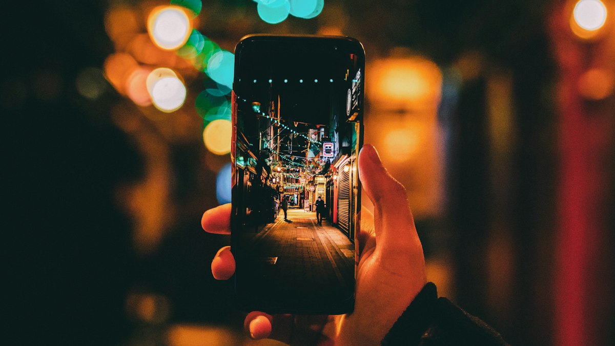 A hand holding a smartphone up to frame a narrow night street lined with lit shop signs in its camera