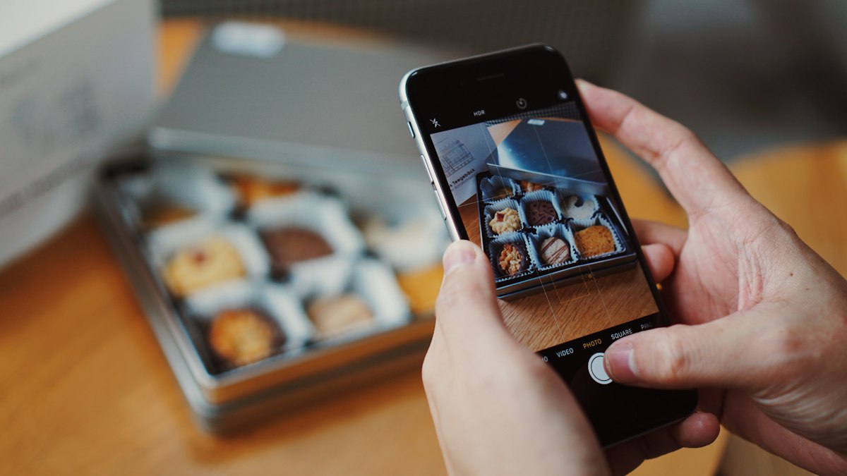 Two hands holding an iPhone in the Camera app, framing a tin of assorted biscuits on a wooden table