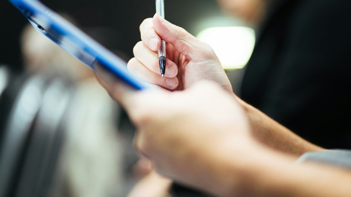 A hand holding a pen over a blue clipboard, with a blurred office in the background