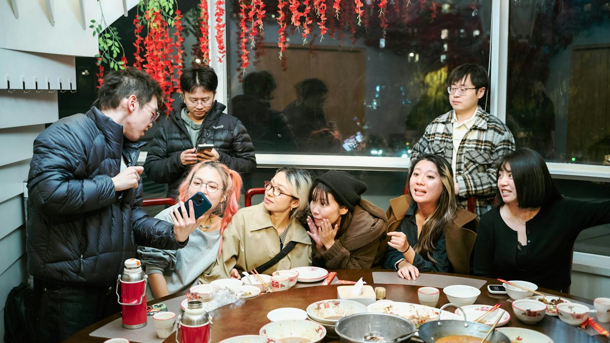 A group of friends around a round restaurant table after a meal, one of them holding up a phone