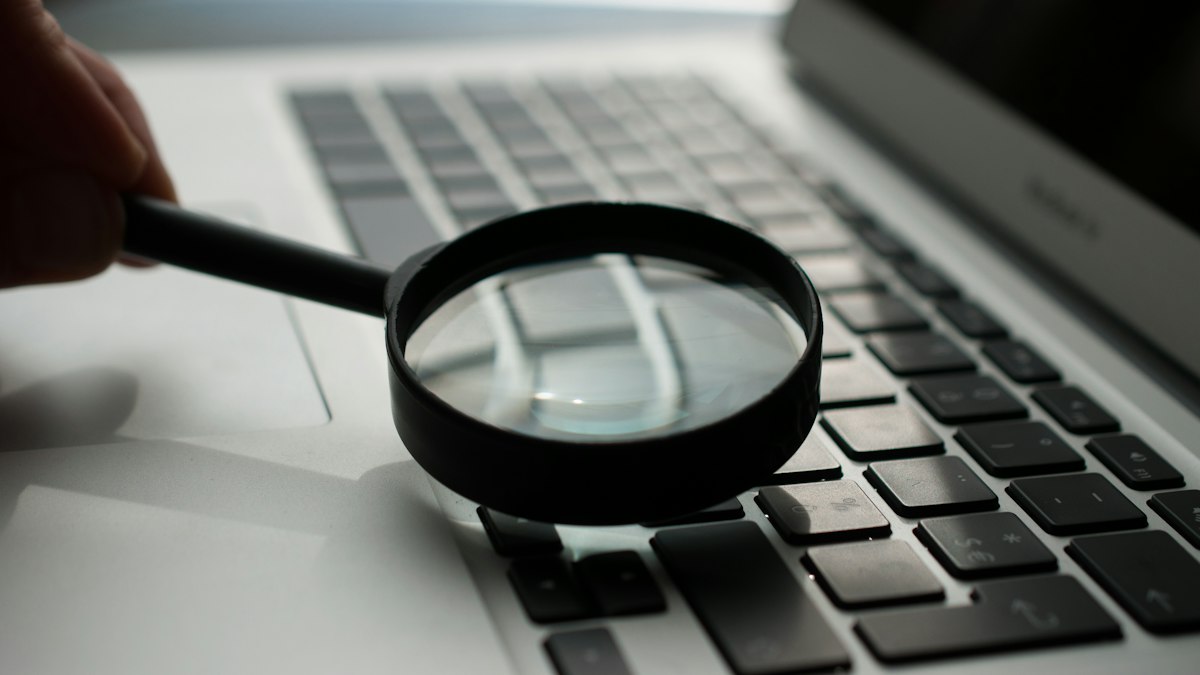 A hand holding a black magnifying glass over the keyboard of a silver laptop